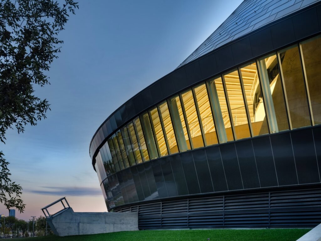 Low-angle exterior shot at dusk showing the sloping glazed curtain wall and metallic cladding of Coronation Park Sports Centre.
