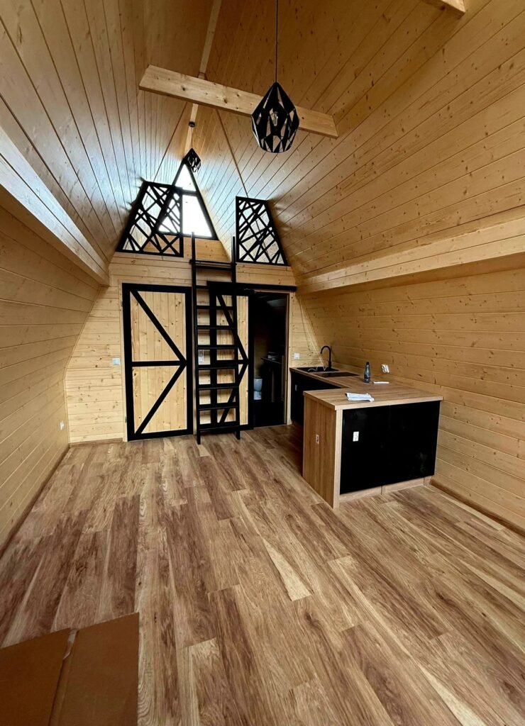 Interior of an A-frame micro cabin featuring light pine wall cladding, wood floors, a compact kitchen island, and a black ladder leading to a sleeping loft.