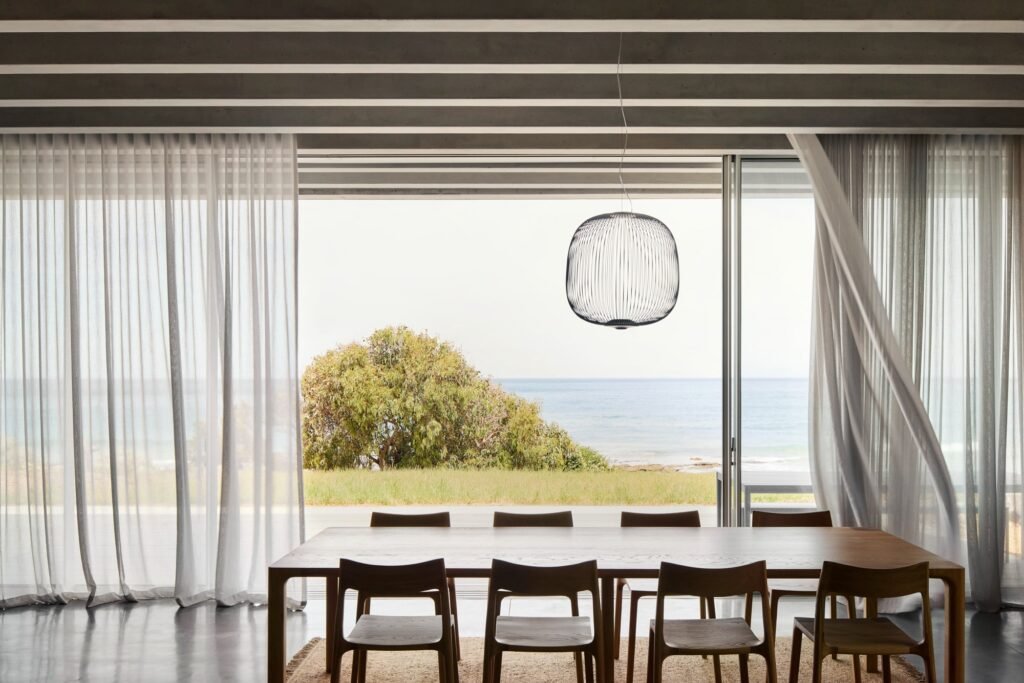 Interior dining space with timber table, sheer curtains, hanging wire pendant, and floor-to-ceiling glass doors opening to ocean view.