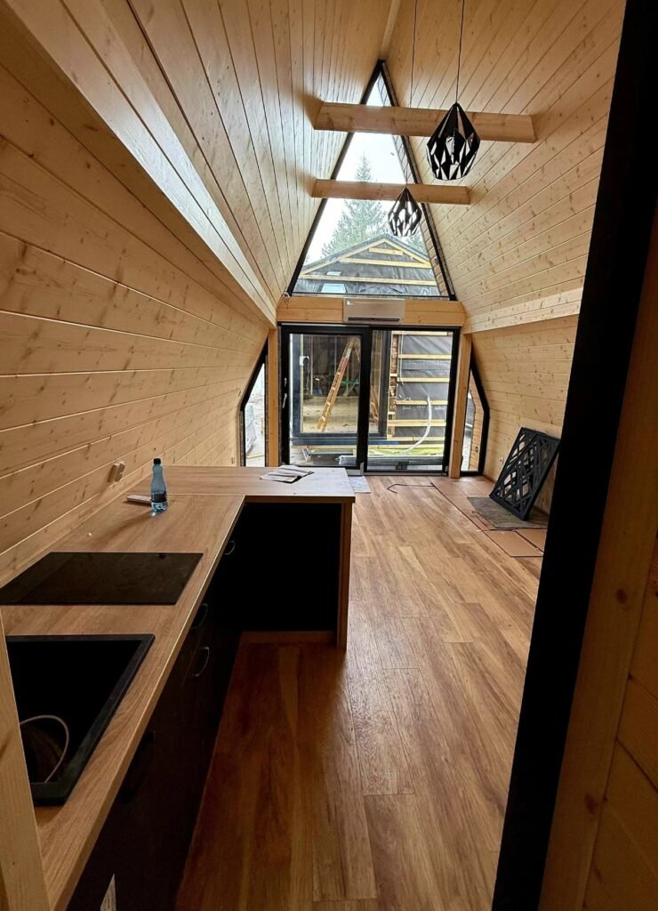 Interior perspective from the kitchen looking toward the large triangular glass entrance of a timber A-frame house.
