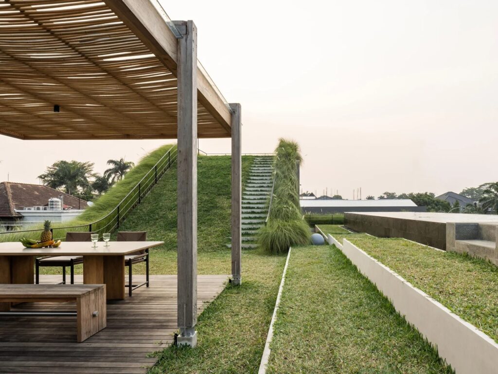 Outdoor dining table under a wooden trellis on a green roof terrace next to grass steps leading upward.