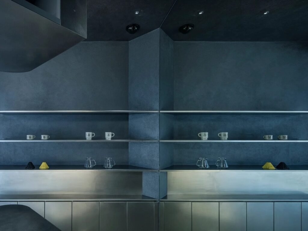 Minimalist dark wall with horizontal stainless steel display shelves holding coffee drippers and mugs.