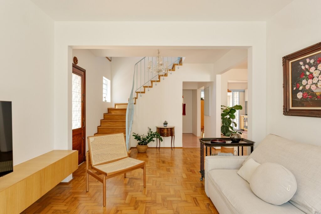 Living room in Casa M showing original Peroba-do-campo wooden floors, a restored staircase with light blue metal railing, and neutral furnishings.