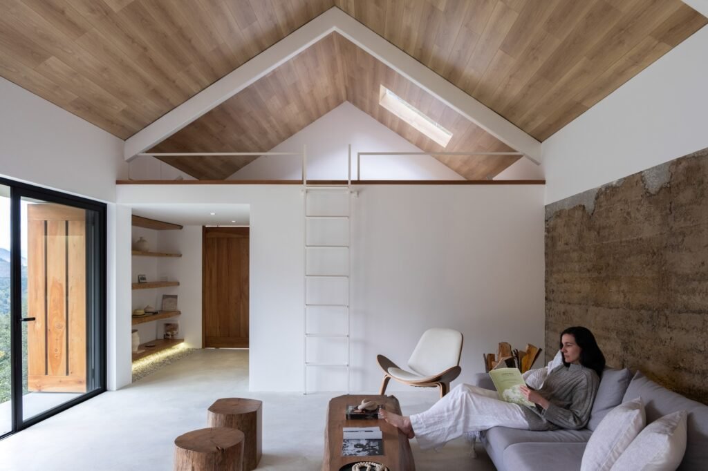 Interior living room featuring an exposed rammed earth wall, light wood vaulted ceiling, white steel mezzanine ladder, and a woman sitting on a sofa reading.