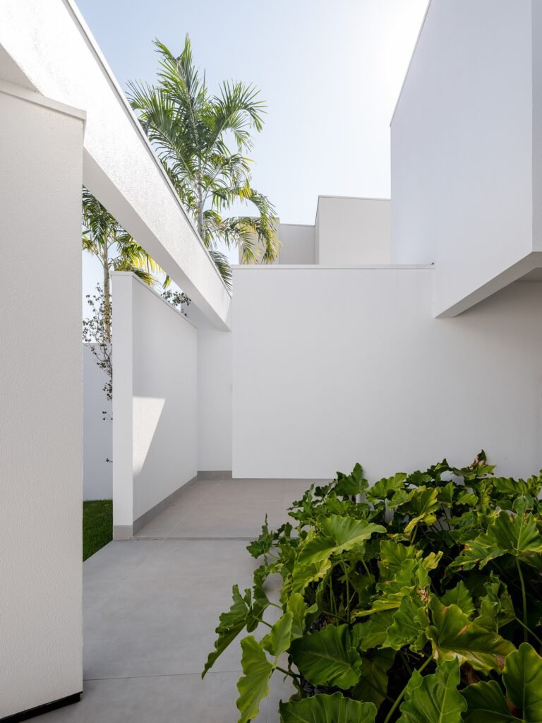 Architectural detail of Sorriso House showing overlapping white walls, a concrete beam overhead, palm trees, and lush green plants, highlighting the contrast of light and shade.