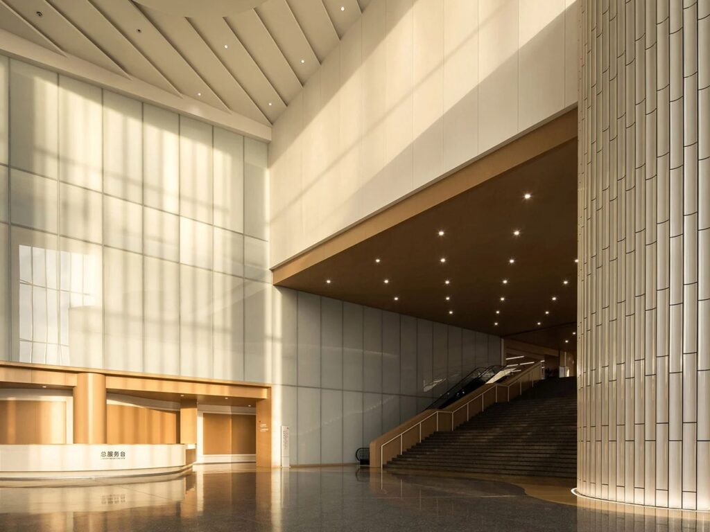 Double-height entrance hall featuring a reception desk, grand staircase, textured cylindrical column, and warm ambient sunlight.