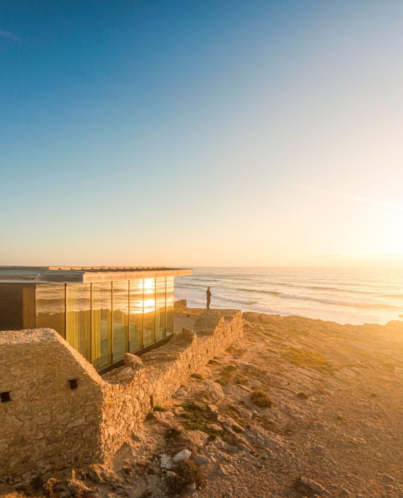 Sunset view of a person standing on the restored stone wall edge of Cresmina Fort next to a modern floor-to-ceiling glass volume facing the ocean.