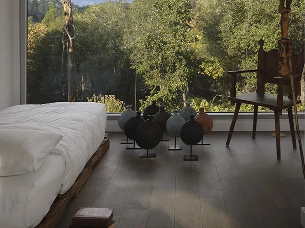 Minimalist bedroom interior with a low platform bed, dark wood flooring, decorative ceramic art objects, and a full-height glass window facing a lush forest.