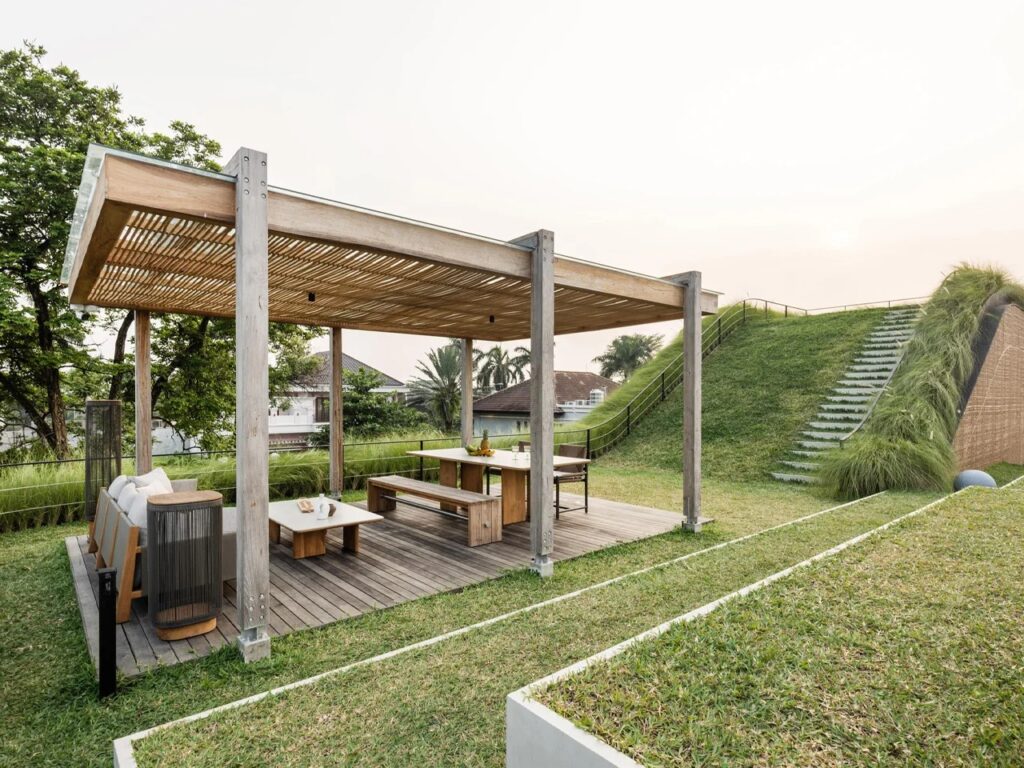 Shaded wooden pergola with outdoor lounge furniture on a green lawn roof terrace next to a grassy slope.