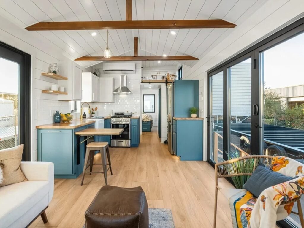 Bright open-plan tiny home interior with white painted wooden walls, exposed ceiling collar beams, blue kitchen cabinets, and large sliding glass patio doors.