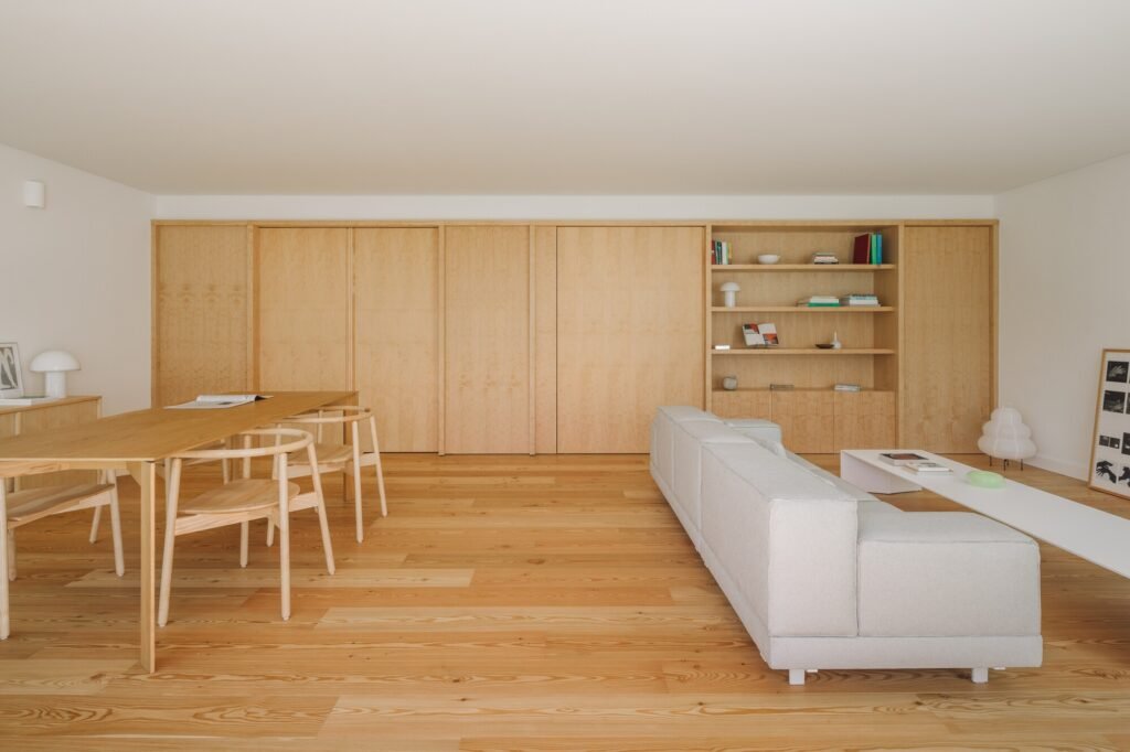 Spacious open-plan living room with light wood floors, a white sofa, a long wooden dining table, and built-in wooden shelving.
