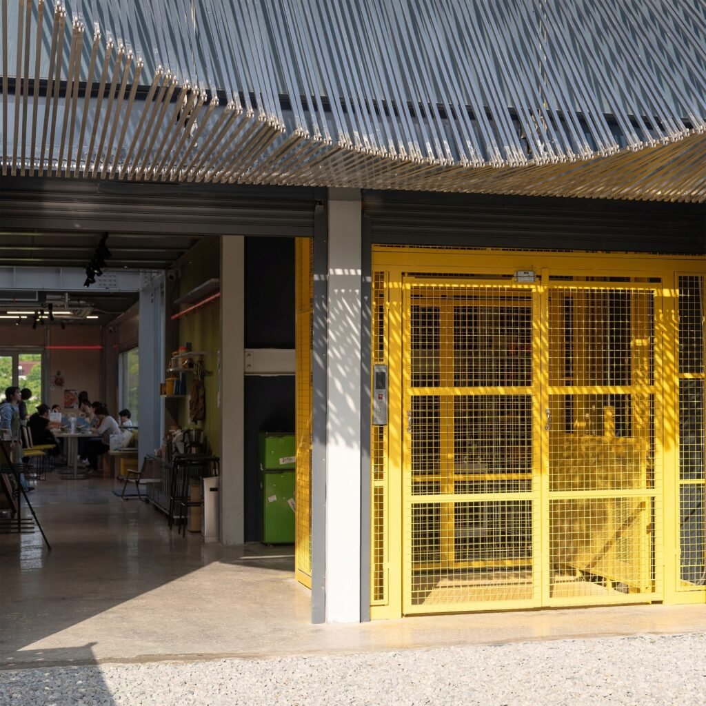 Industrial yellow mechanical hoist cage gate inside Spitfire Roastery alongside a slatted canopy and dining area.