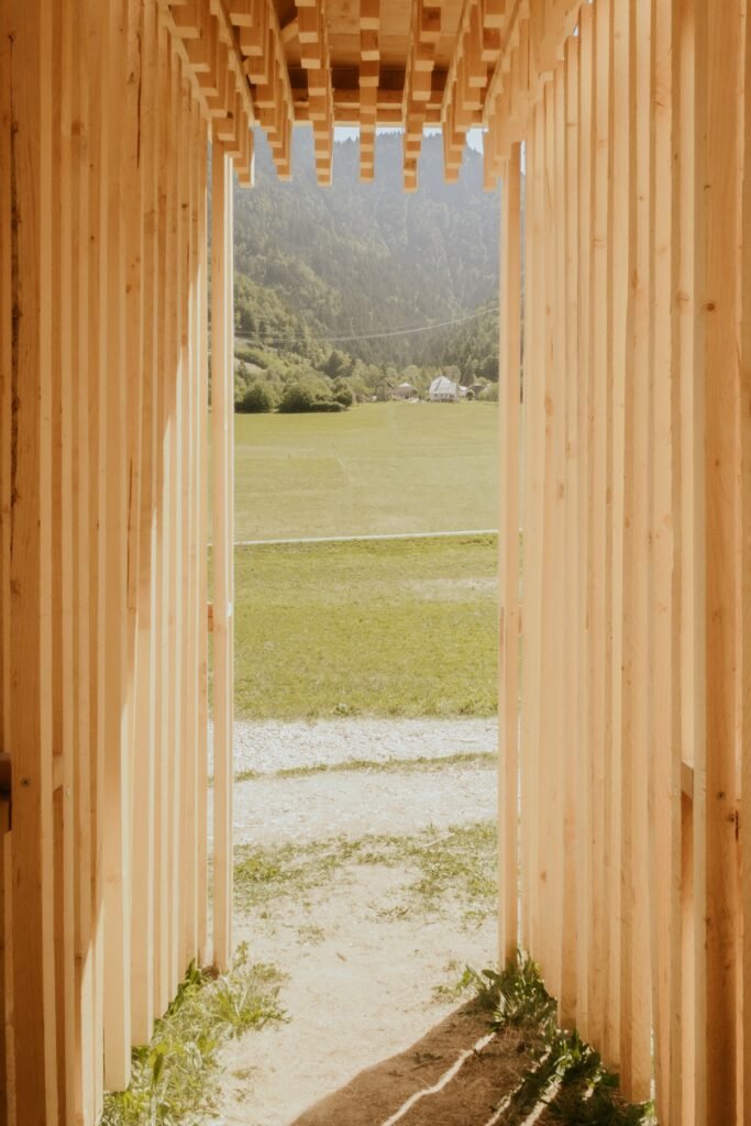 View from inside a wooden slat corridor framing a scenic landscape view of a green meadow and mountain forest.