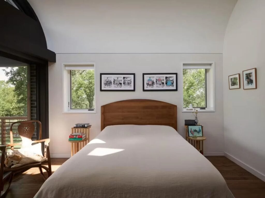 Bright bedroom in Crescendo house with white walls, wooden headboard bed, and large square windows overlooking trees.