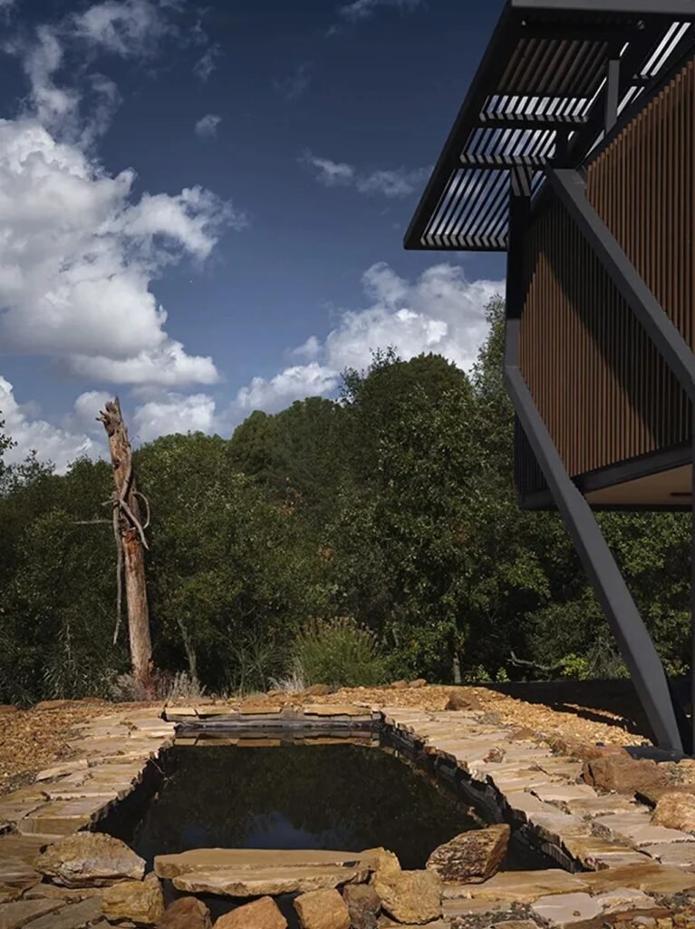 Low-angle view of a natural stone-lined reflective pool near the steel and timber structure of Null House under a blue cloudy sky.