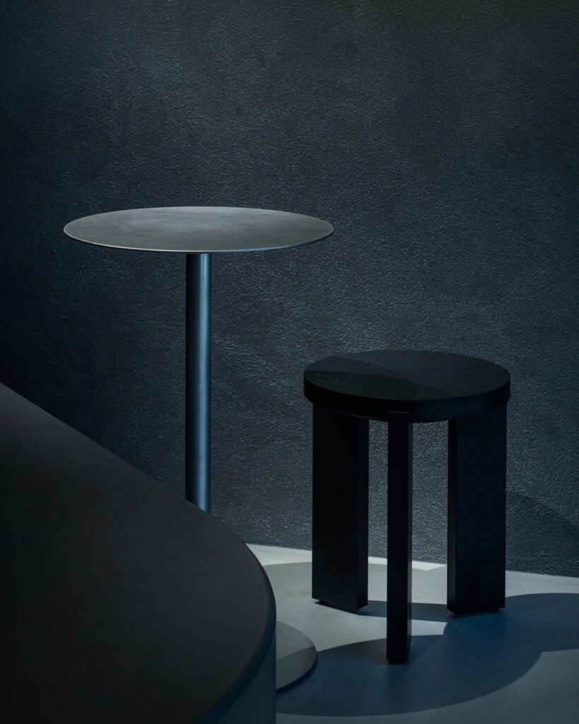 Minimalist round metal coffee table and black wooden tripod stool against dark textured wall.