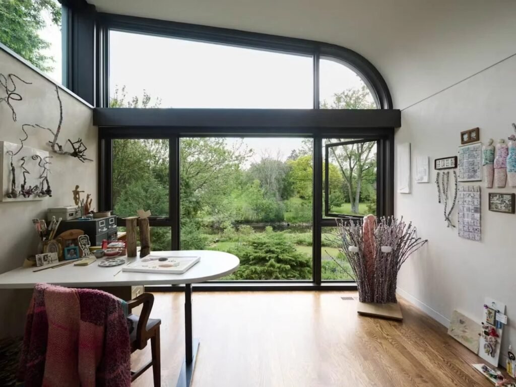 Sunlit art studio room with working desk, art supplies, and large curved transom window facing leafy outdoors.