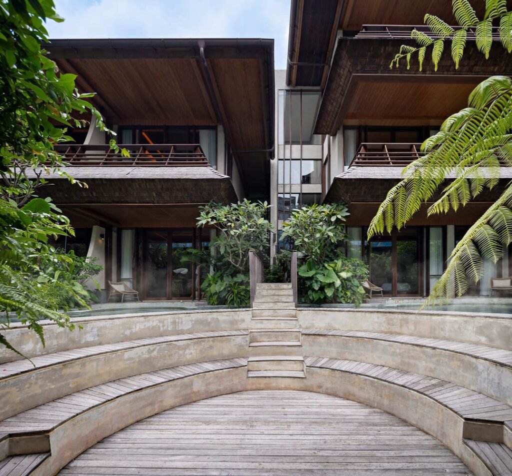 Curved organic swimming pool and sunken wooden amphitheater steps surrounded by lush tropical ferns and resort villas.