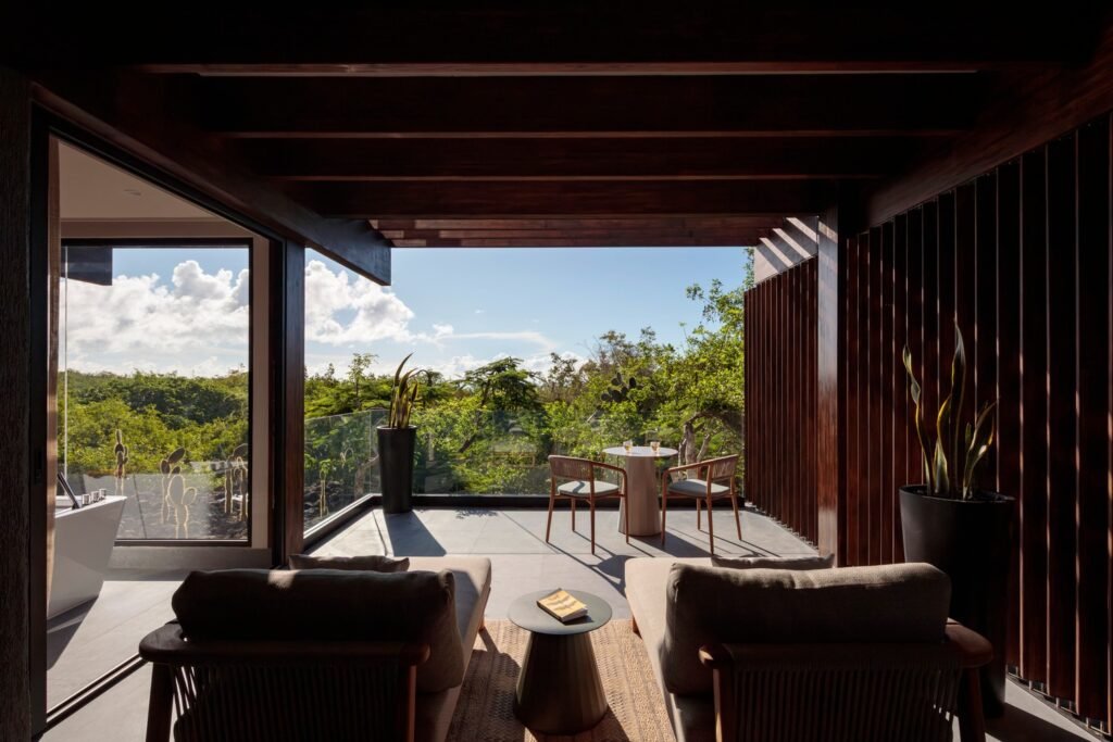 View from a modern hotel room interior looking out to a spacious, covered wooden balcony. Two wooden lounge chairs and a small table are arranged on the gray tiled balcony, with clear glass railings offering panoramic views of the mangrove forest and distant sky.