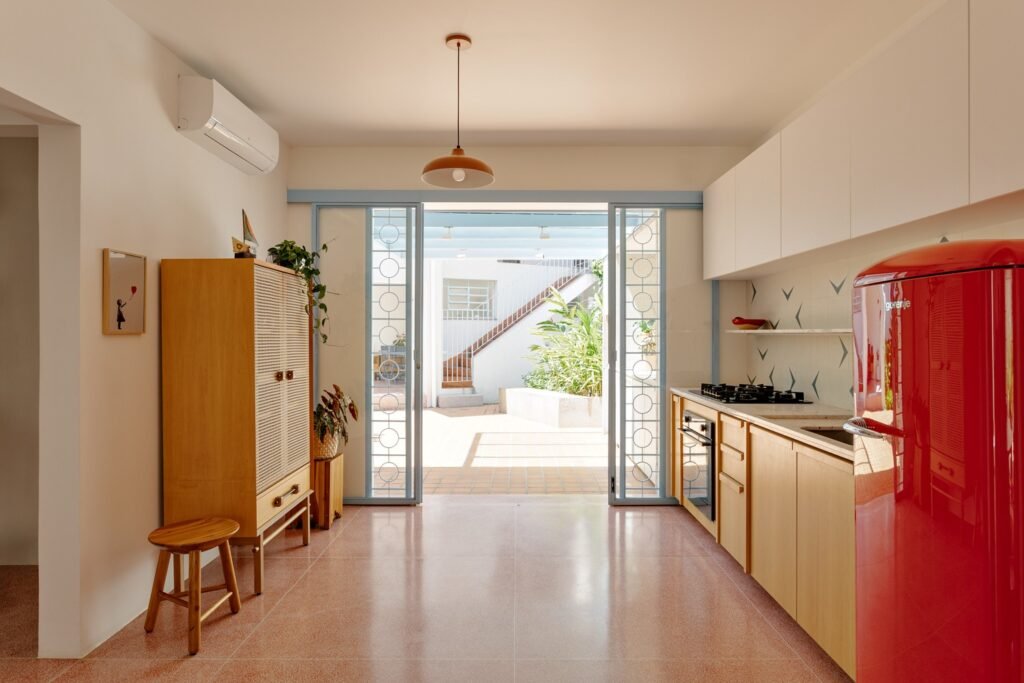 Modern kitchen area with custom pink terrazzo flooring, light wood cabinetry, a red retro refrigerator, and glass doors opening to the outdoor courtyard.