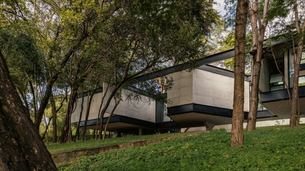 Exterior view of concrete masses and steel beams supported on sloped ground amid trees.