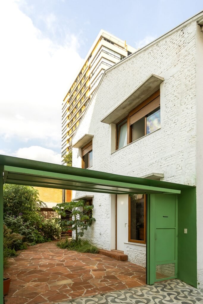 White painted brick exterior facade of Casa em São Paulo with a green metal entrance canopy and sandstone paved driveway.