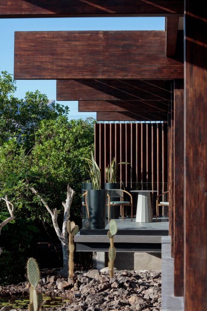 Detailed close-up focusing on the prominent dark laminated wood beams, roof overhangs, and vertical wooden louvers of a Finch Bay Hotel balcony. Potted snake plants and furniture are visible.