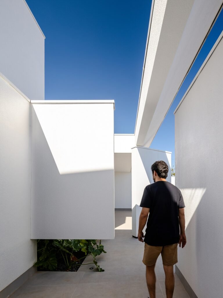 A man walking along an outdoor walkway between stark white geometric volumes and a raised wall at Sorriso House under a bright blue sky.