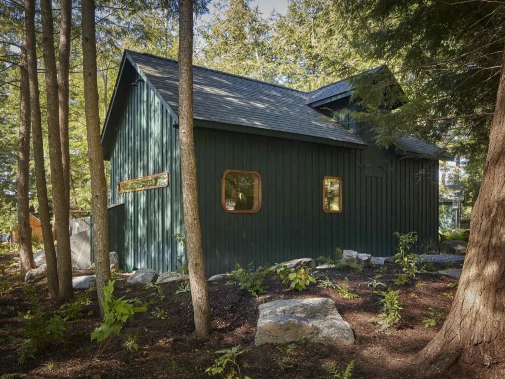 Dark green exterior of transformed historic icehouse cabin with pitched roof nestled in Muskoka pine forest.