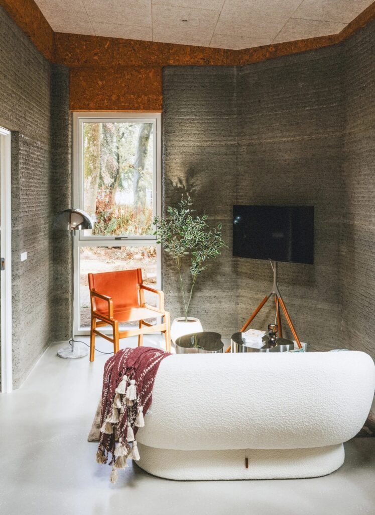 Sunlit interior living room in a student apartment with exposed 3D printed concrete walls, modern couch, and window views.