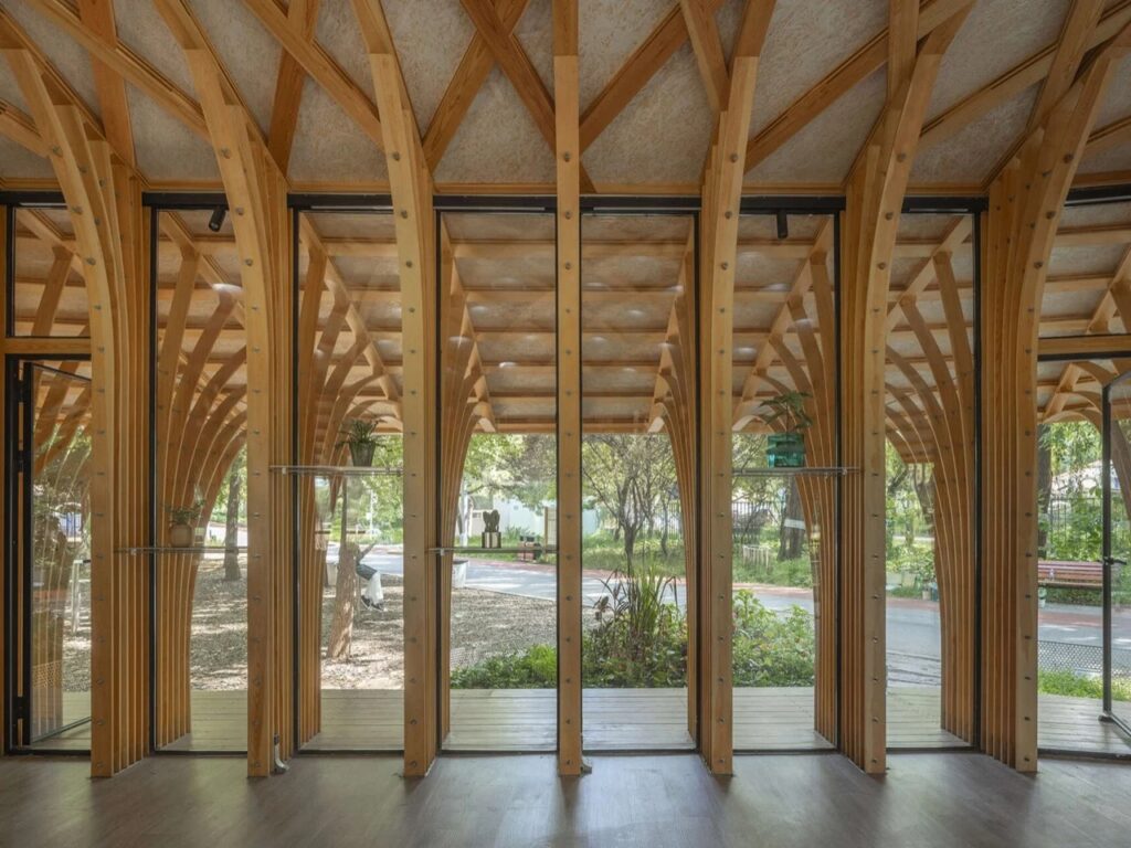 Interior view looking through full-height glass windows between structural wood columns out toward the surrounding park trees.