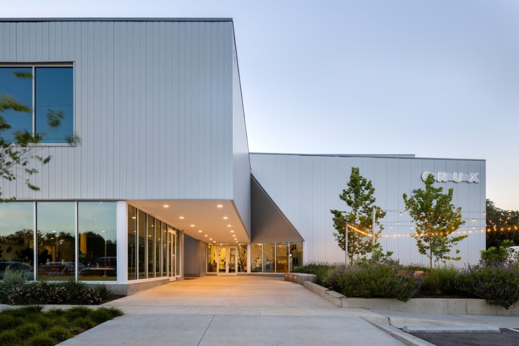 Main entrance plaza of Crux South illuminated at dusk featuring glazed storefronts and elevated signage.