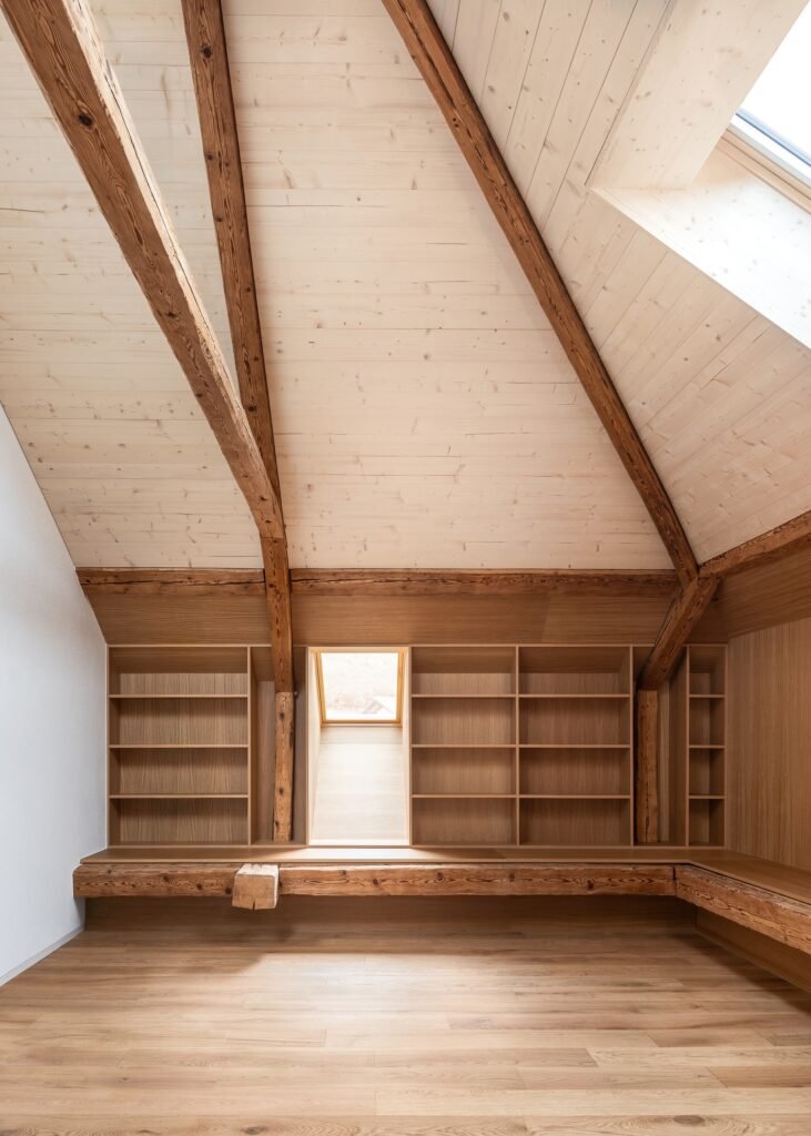 Attic interior with sloping light wood ceiling, dark exposed historic timber rafters, skylight, light wood flooring, and built-in wall-to-wall shelving.