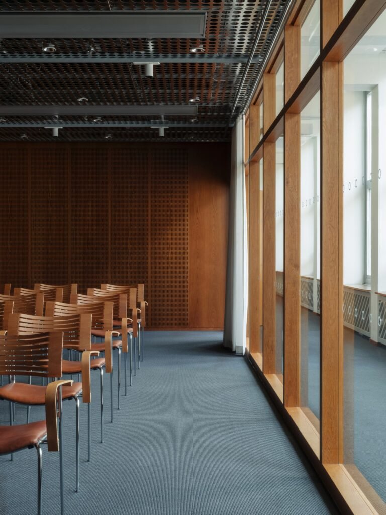 Modern conference room at Folkets Hus featuring bentwood chairs, blue carpet, perforated metal ceiling, and large wooden-framed windows.