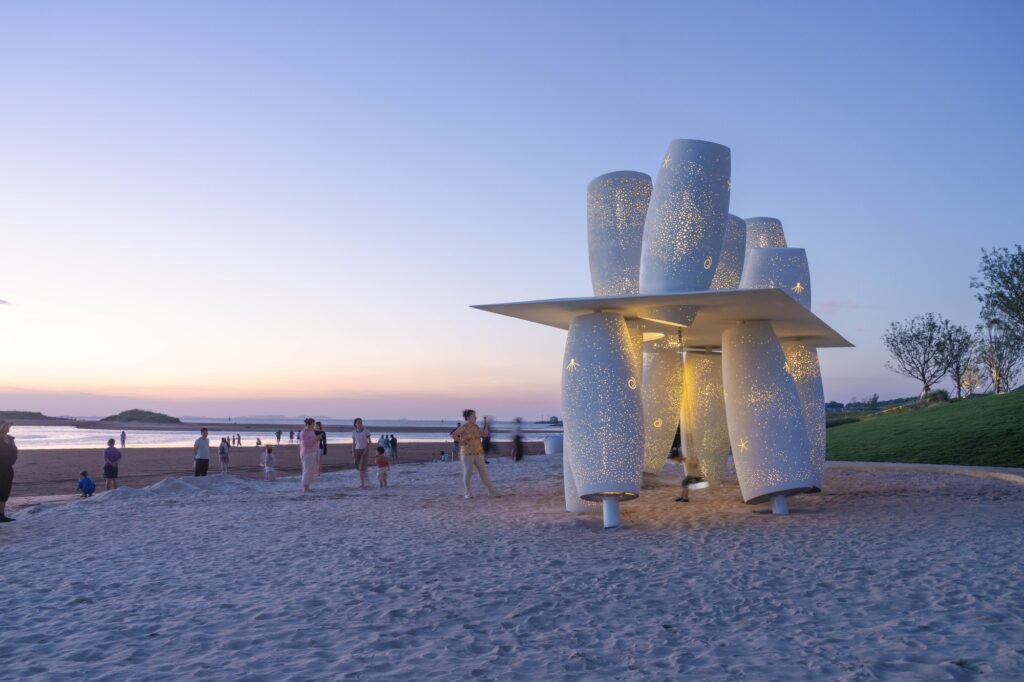 Evening view of the Coral installation on the beach, illuminated from within through thousands of tiny perforations against a sunset sky.