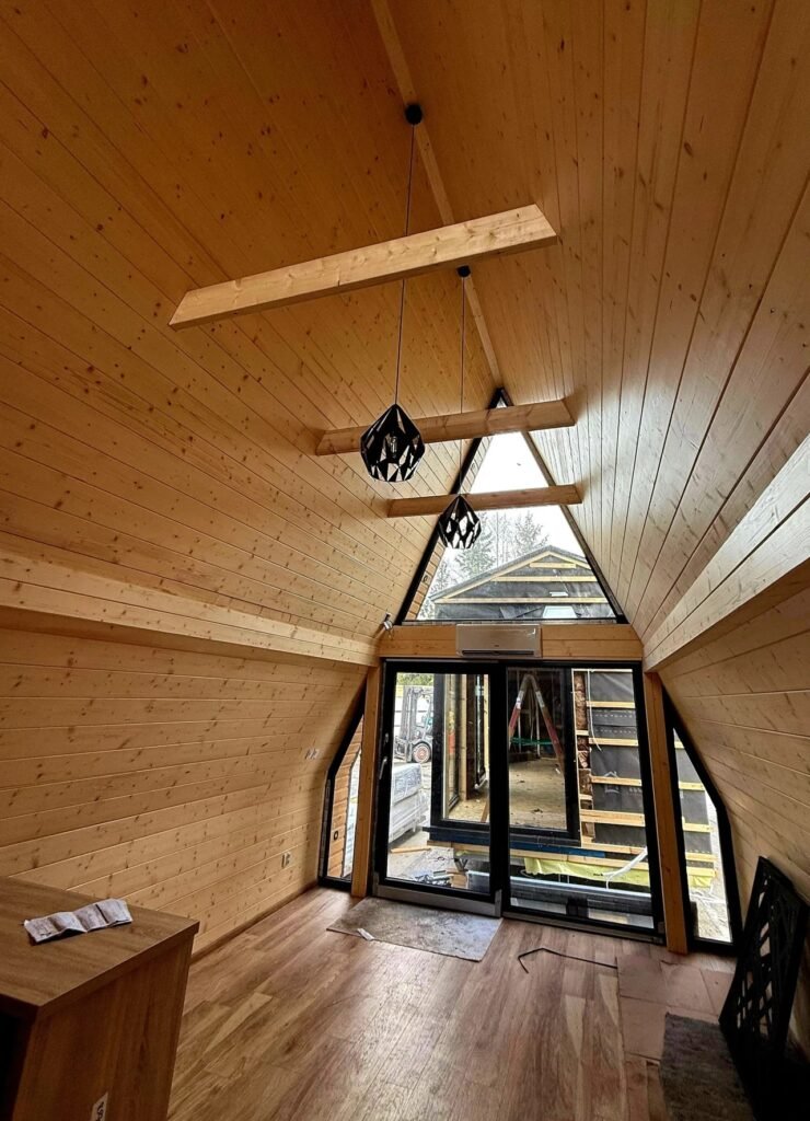 Looking up at the angled pine ceiling of an A-frame cabin with exposed crossbeams and black geometric pendant lights hanging near a top window.