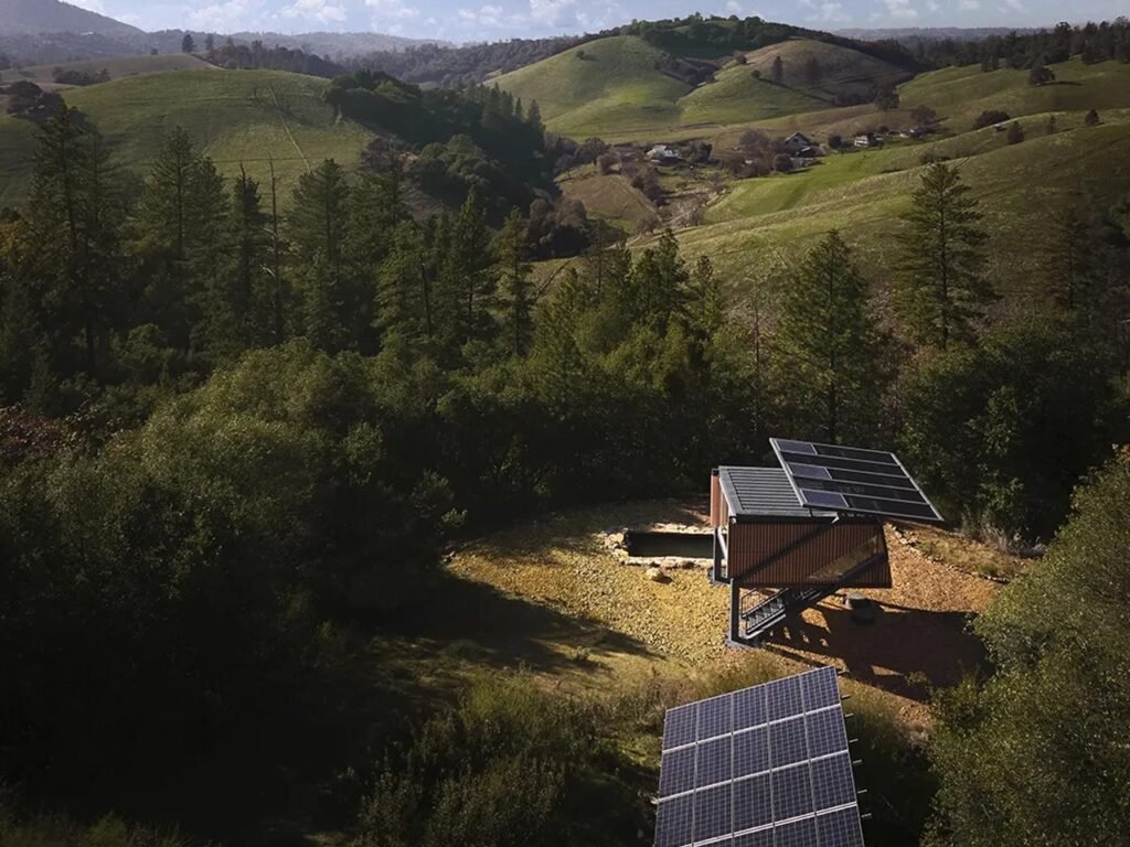 Aerial drone perspective of Null House situated on a grassy hill with photovoltaic solar arrays mounted on the roof and ground nearby.