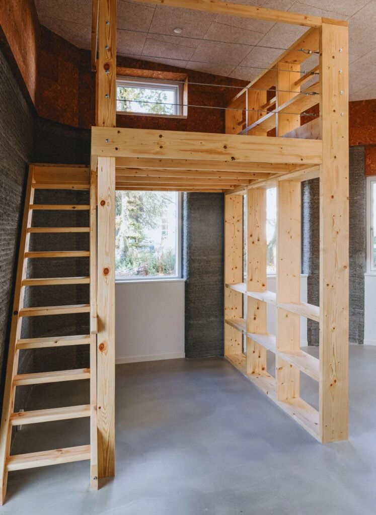 Built-in wooden loft bed structure with ladder inside a student unit with exposed 3D printed concrete walls.