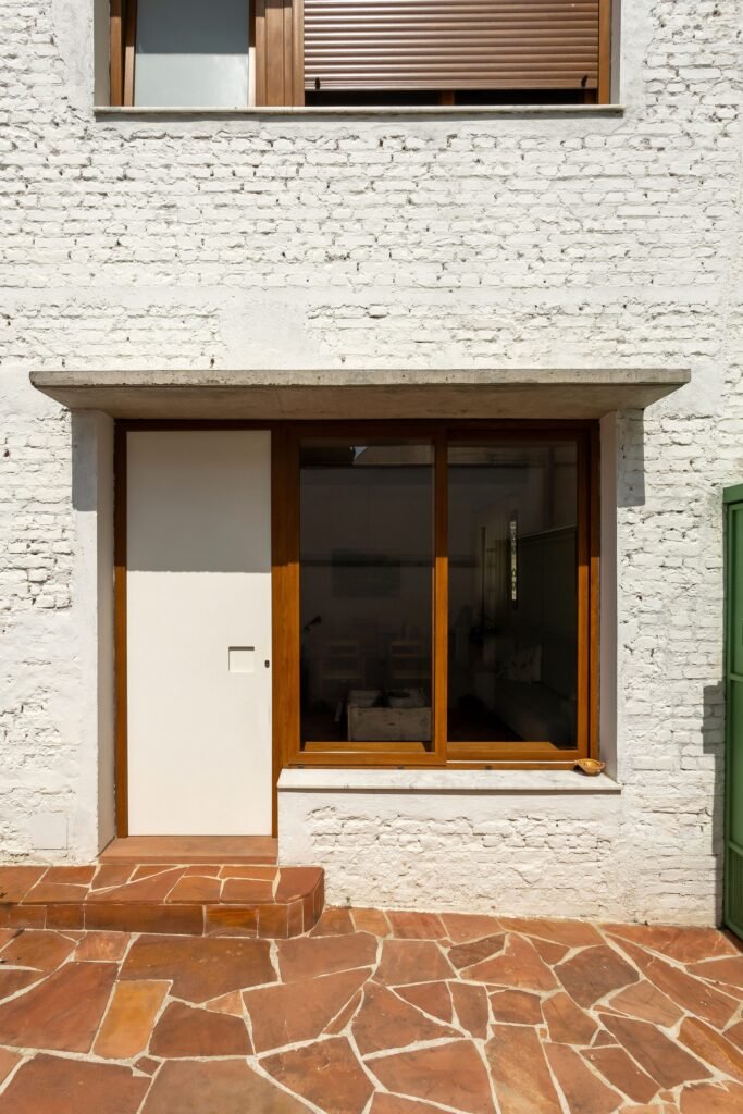 Ground level entry door with wood frame sliding glass window under a concrete cantilevered overhang and sandstone tile paving.