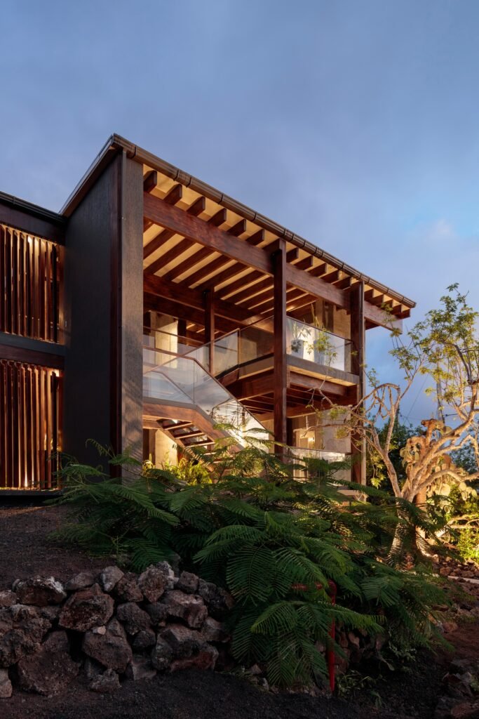 A two-story glass-and-wood main building of the Finch Bay Hotel illuminated from within at twilight, with warm lights highlighting an internal staircase. Lush green vegetation and rocks fill the foreground under a dusky blue sky.