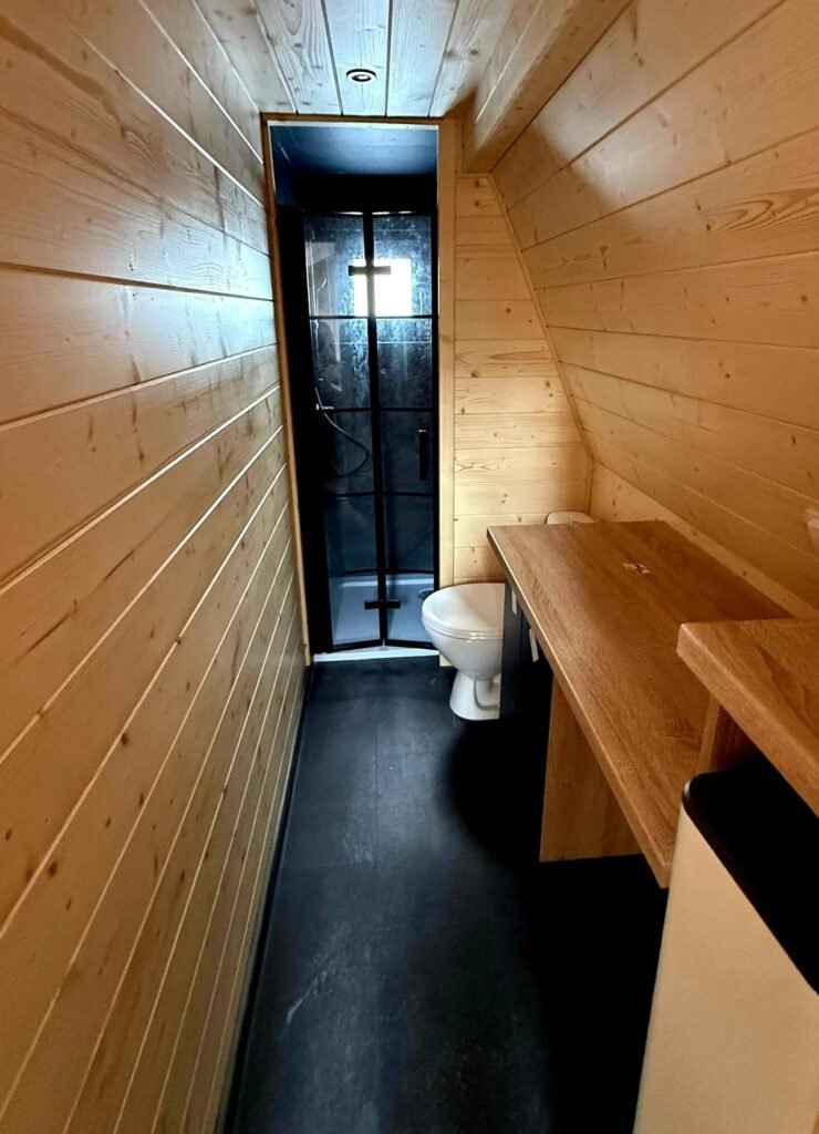Narrow bathroom interior with pine plank walls, dark flooring, wooden vanity counter, toilet, and black-framed glass shower stall at the end.