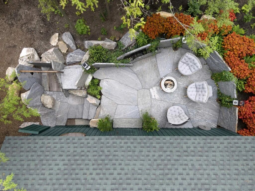 Top-down aerial view of stone patio, fire pit, outdoor shower, and roof of green spa cabin.