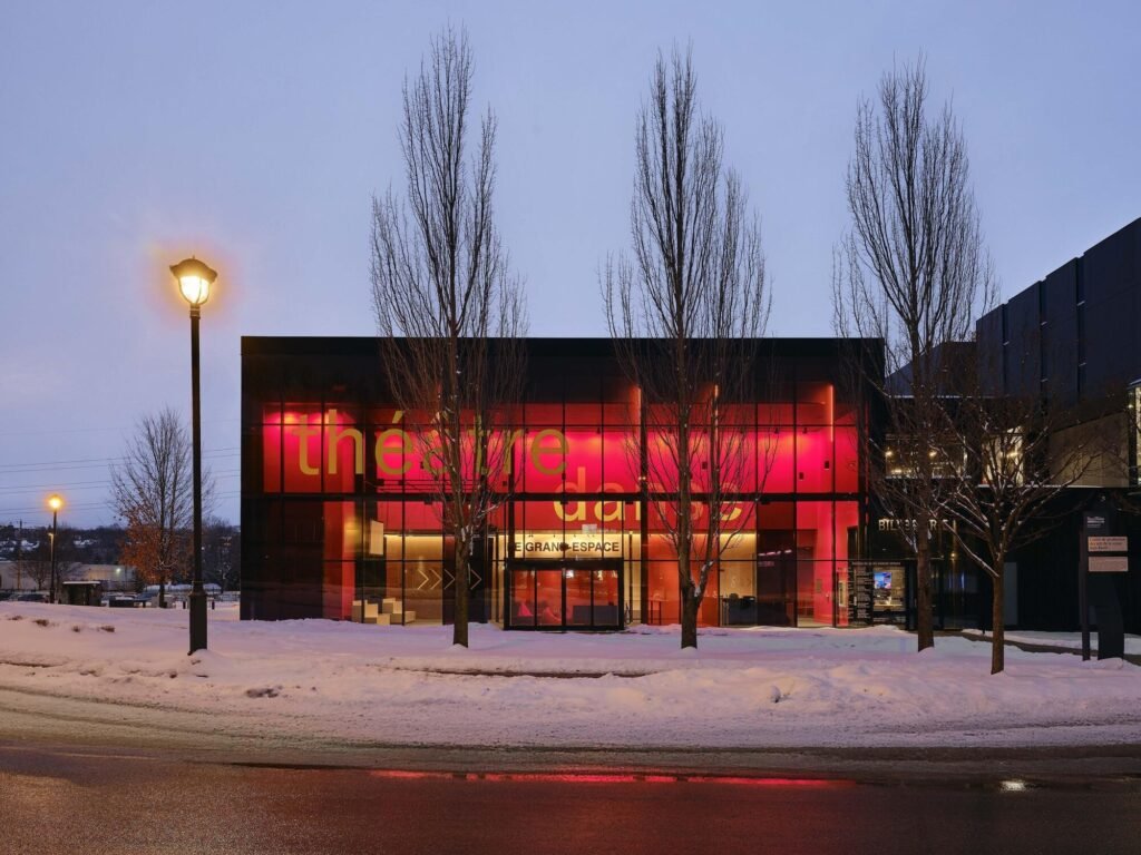 Night exterior elevation of Le Grand-Espace glowing with warm red and pink interior lights behind bare trees in winter.
