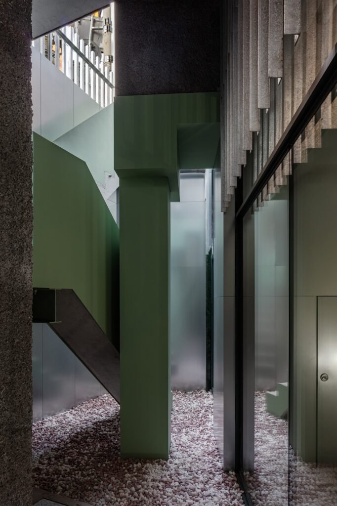 Narrow circulation passage showing contrasting green structural element, rough concrete wall, and metallic panels.