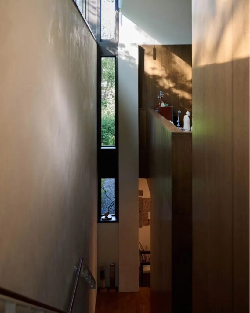 Narrow double-height interior lightwell and stairwell in Crescendo home with tall slot windows and natural wood wall panels.