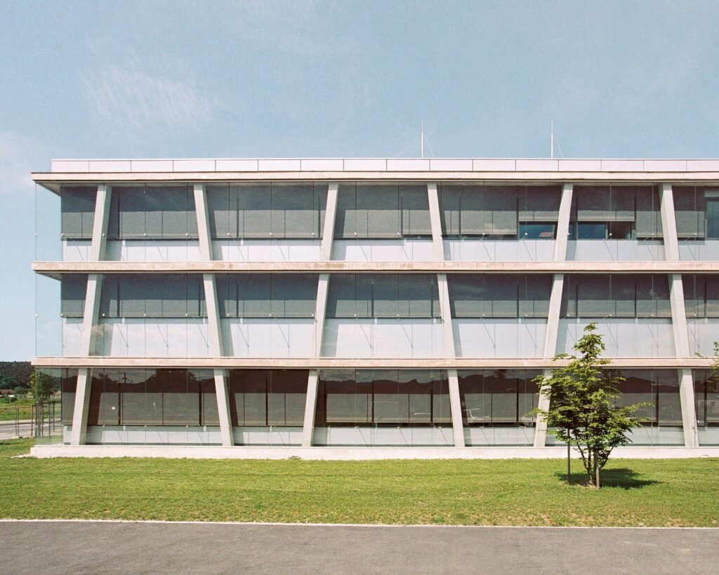 Straight-on architectural photo of the three-story glass and concrete facade featuring inclined perimeter columns and integrated sunshades.
