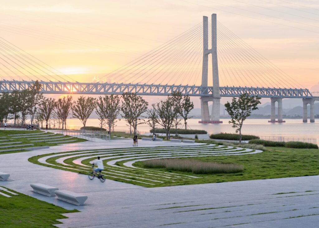 A cyclist riding along a curved concrete path surrounded by sculpted grass terraces at sunset, with the cable-stayed bridge lit up by the setting sun.