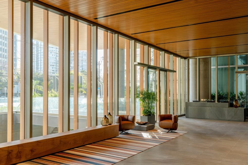 Interior view of Jer&ocirc;nimo Faria Lima main lobby featuring warm wooden slatted ceiling, floor-to-ceiling glass curtain wall, reception desk, and designer seating.