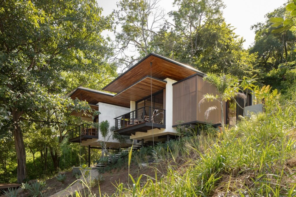 Low-angle view of elevated tropical villa modules supported by black steel columns along a steep hillside.