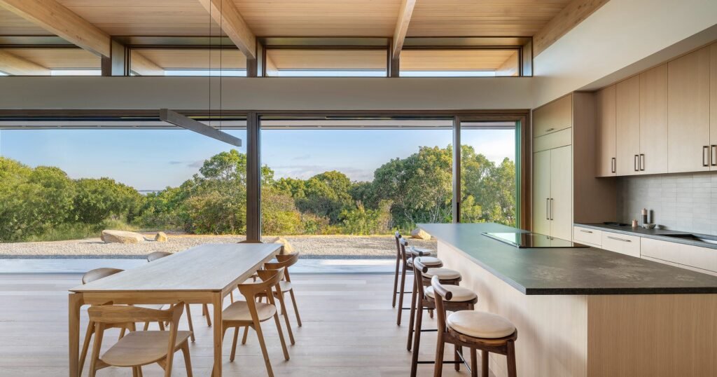 Interior of Veil House displaying a light wood dining table, kitchen island, and massive glass windows overlooking the forest foliage.